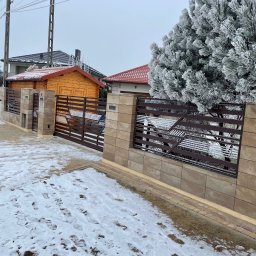 Dawid Karolewski - Zimowy krajobraz z brązową bramą wjazdową i ogrodzeniem z beżowych bloczków. Na pierwszym planie ślady na śniegu, w tle ośnieżone drzewo i budynek.