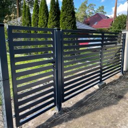 Dawid Karolewski - Nowoczesna, grafitowa brama wjazdowa z poziomymi szczeblami, częściowo otwarta, z widocznym fragmentem ogrodzenia z siatki i zieleni w tle.