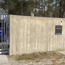 Dawid Karolewski - Nowoczesna brama wjazdowa z białymi, pionowymi panelami, betonowy mur z numerem domu '2' i skrzynką na listy, widok na zaparkowany samochód za bramą.