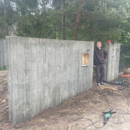 Dawid Karolewski - Betonowy mur ogrodzeniowy z otworem na skrzynkę, obok którego stoi uśmiechnięty mężczyzna. Na ziemi narzędzia budowlane. W tle drzewa.