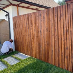 Dawid Karolewski - Montaż drewnianego ogrodzenia panelowego z pionowych desek. Widoczny fragment domu i trawnik z kamiennymi płytami. Fachowiec w trakcie pracy.