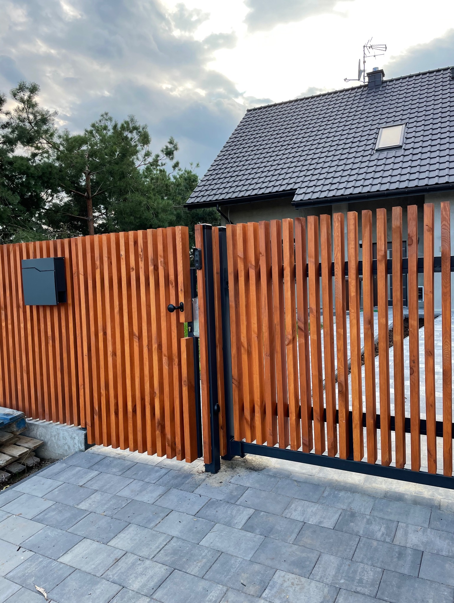 Nowoczesna brama wjazdowa z drewnianymi lamelami w ciepłym odcieniu brązu, czarna skrzynka na listy, szara kostka brukowa i dom w tle pod pochmurnym niebem.