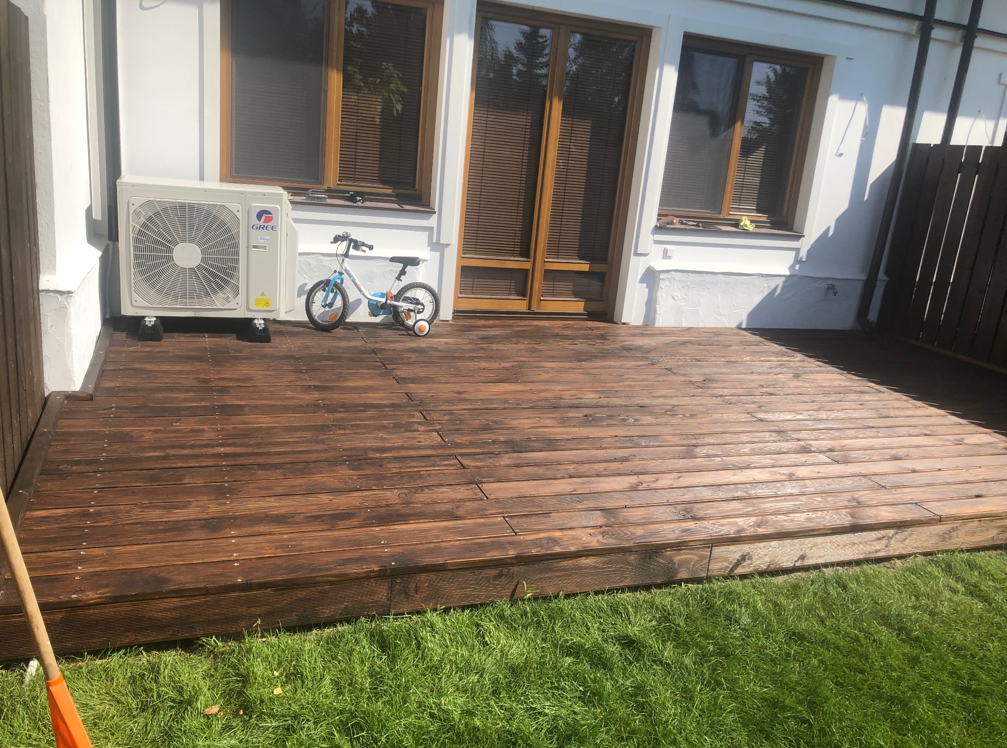 Nowy, drewniany taras przy domu z klimatyzatorem Gree i rowerkiem dziecięcym. Świeżo położone deski tarasowe, widok na trawnik.