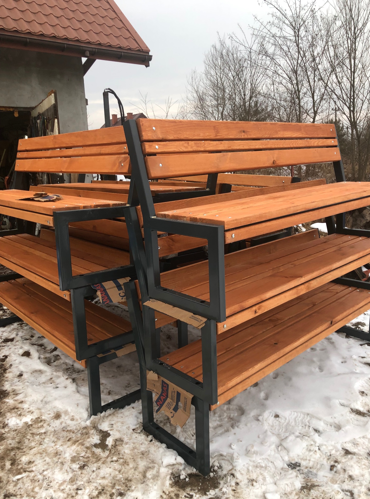 Stos kilku gotowych, drewniano-metalowych ławek ogrodowych na śniegu, z widocznym budynkiem w tle. Konstrukcja z profili, drewno w ciepłym odcieniu brązu.