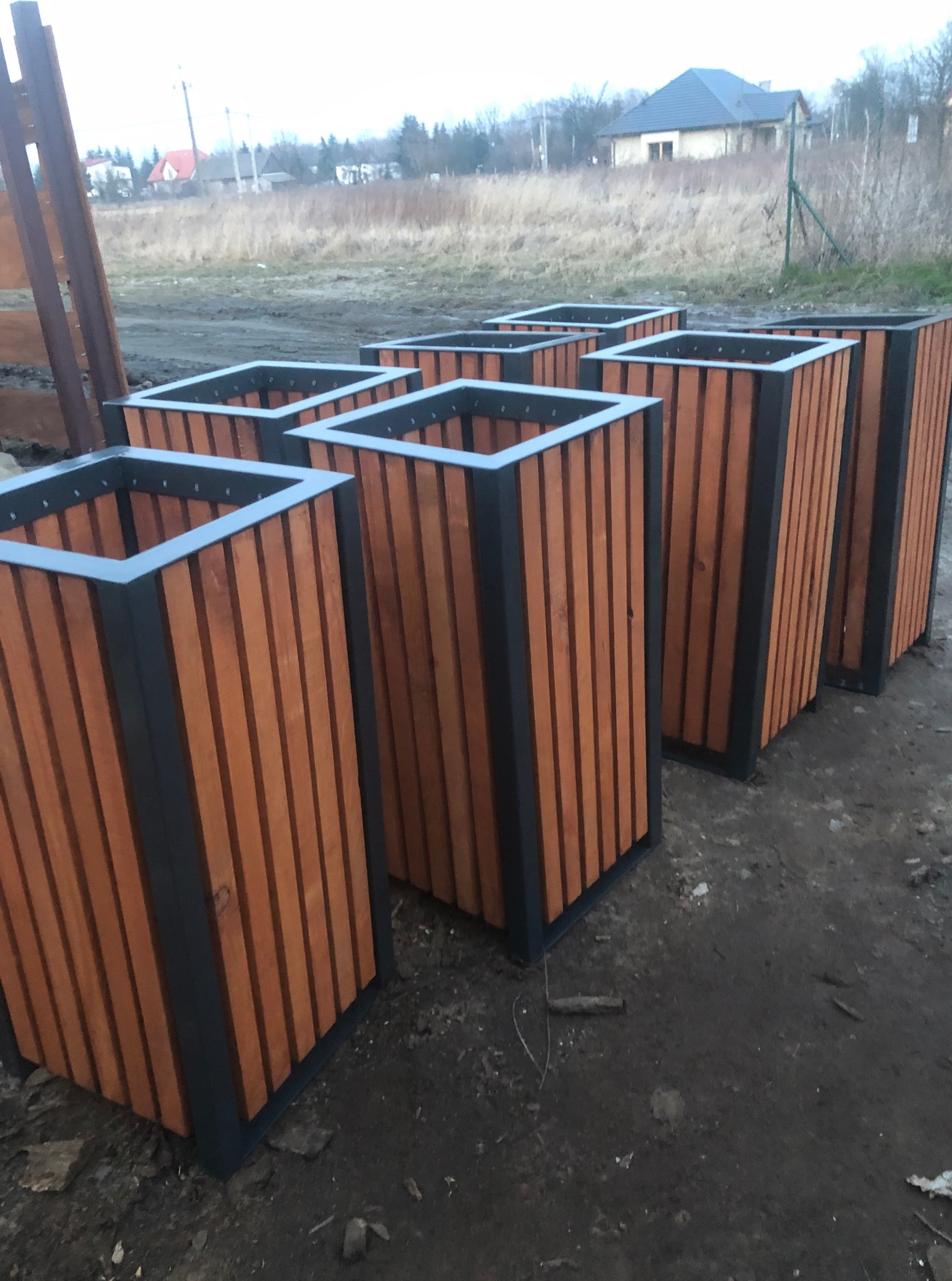 Rząd drewniano-metalowych donic ogrodowych w kształcie prostopadłościanu, ustawionych na ziemi. W tle widoczny dom i drzewa.