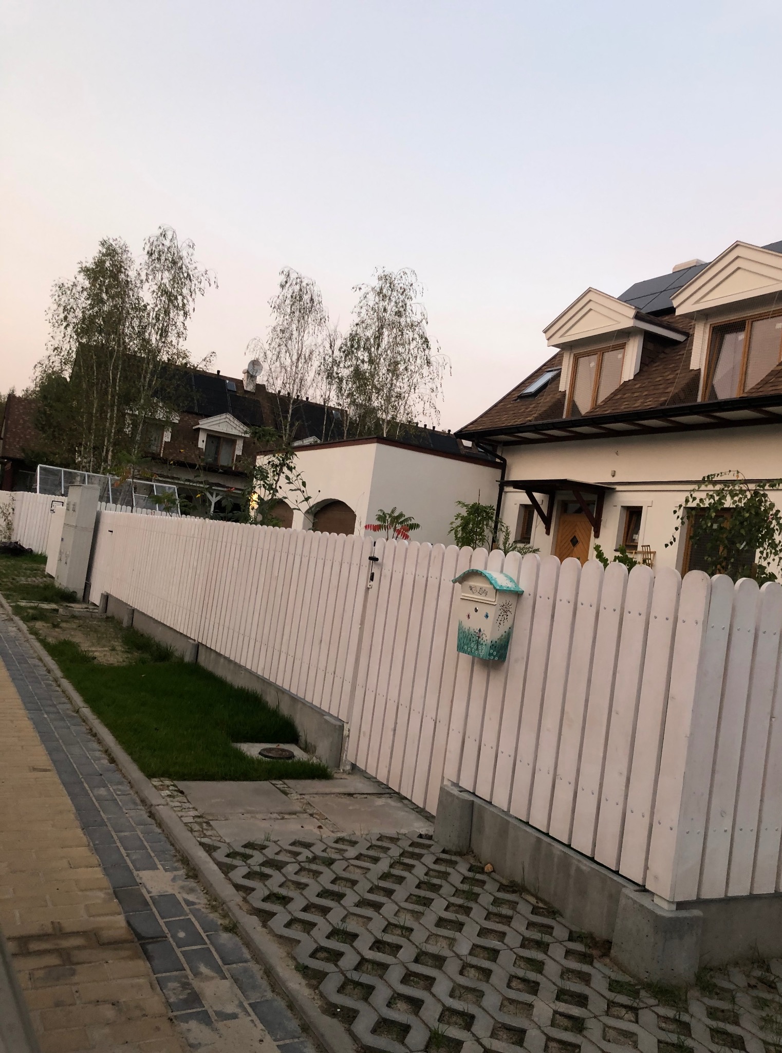 Białe, drewniane ogrodzenie posesji z ozdobną skrzynką na listy. Widok z chodnika na dom z brązowym dachem i oknami na poddaszu. Trawa przy ogrodzeniu.