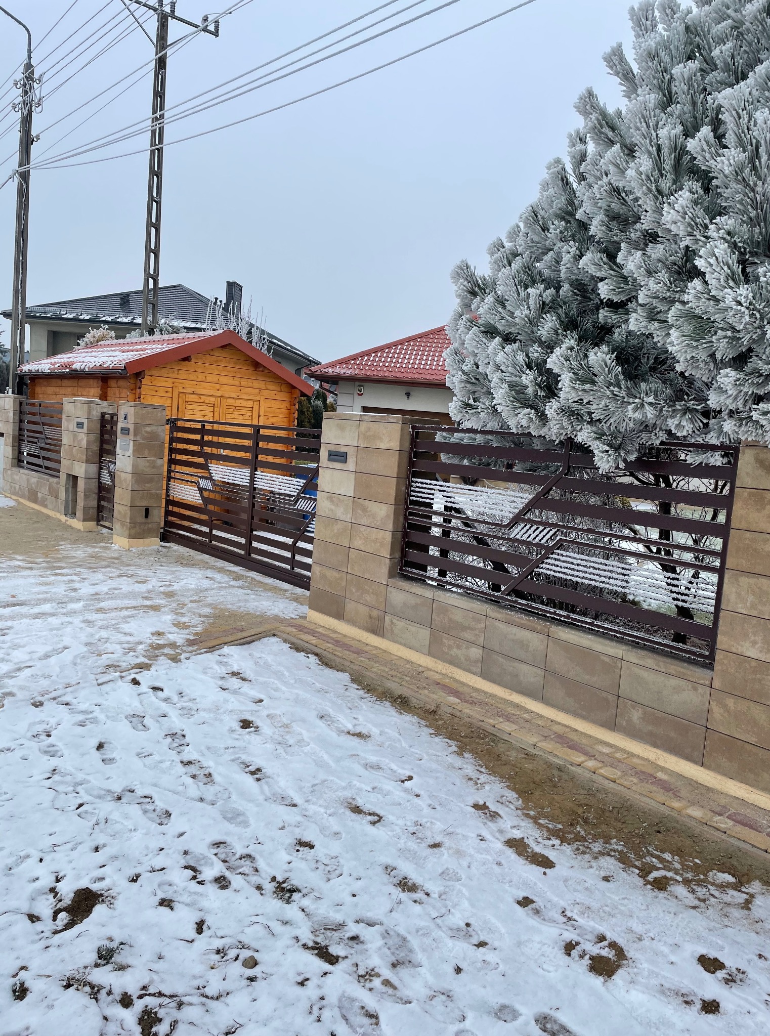 Zimowy krajobraz z brązową bramą wjazdową i ogrodzeniem z beżowych bloczków. Na pierwszym planie ślady na śniegu, w tle ośnieżone drzewo i budynek.