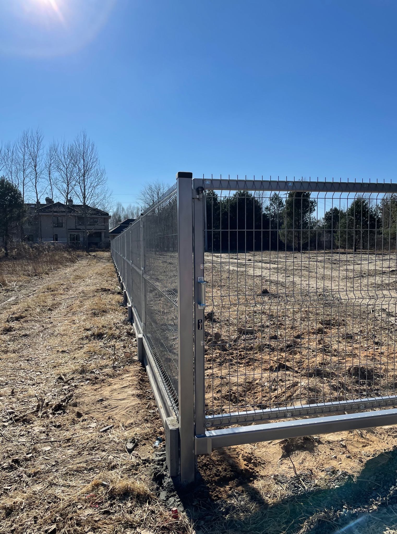 Nowoczesne ogrodzenie panelowe z metalową siatką, widok z perspektywy ziemi, z domem w tle. Słoneczny dzień, błękitne niebo. Minimalistyczny design.