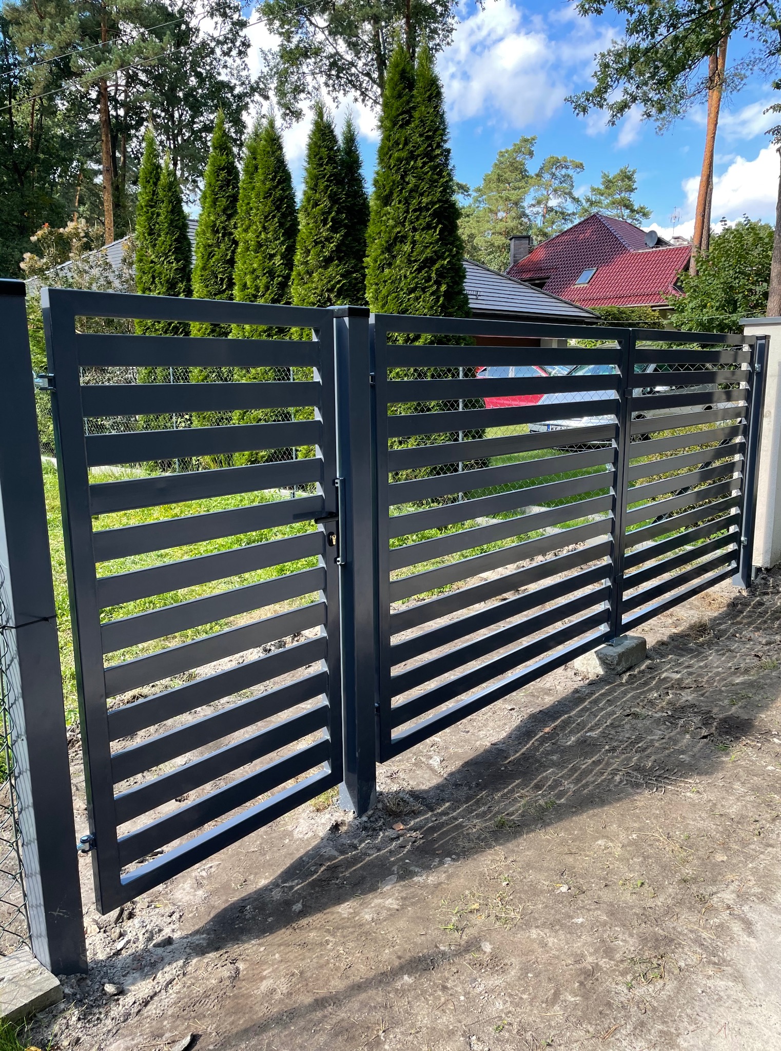 Nowoczesna, grafitowa brama wjazdowa z poziomymi szczeblami, częściowo otwarta, z widocznym fragmentem ogrodzenia z siatki i zieleni w tle.