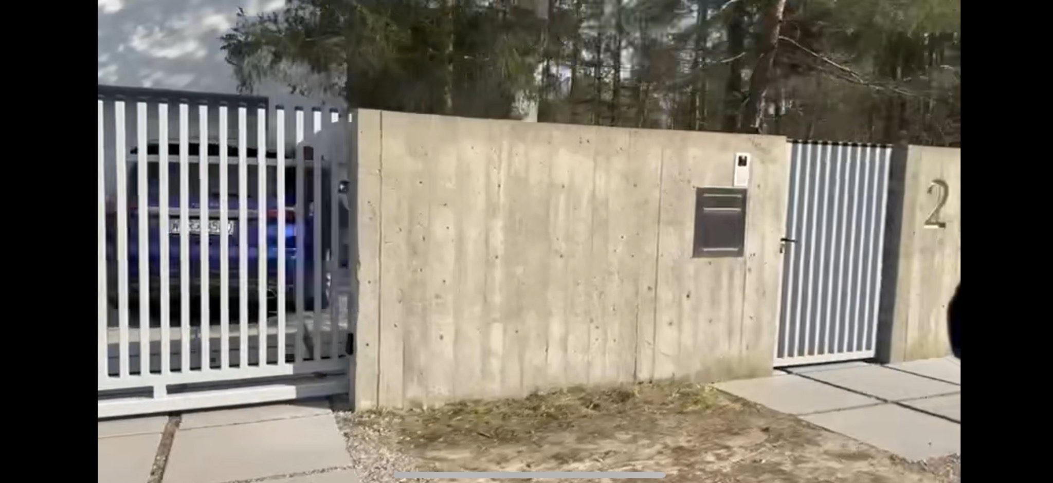 Nowoczesna brama wjazdowa z białymi, pionowymi panelami, betonowy mur z numerem domu '2' i skrzynką na listy, widok na zaparkowany samochód za bramą.