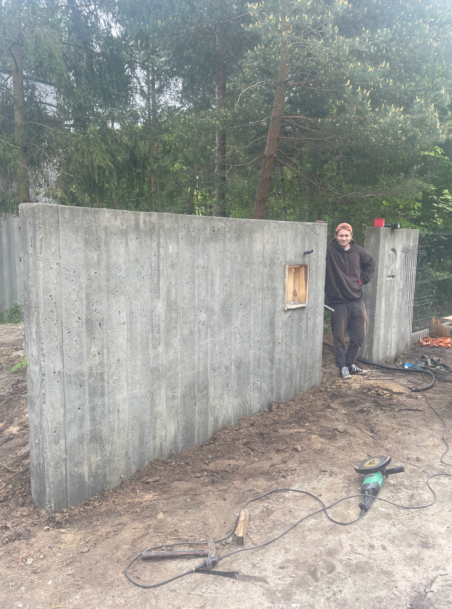 Betonowy mur ogrodzeniowy z otworem na skrzynkę, obok którego stoi uśmiechnięty mężczyzna. Na ziemi narzędzia budowlane. W tle drzewa.