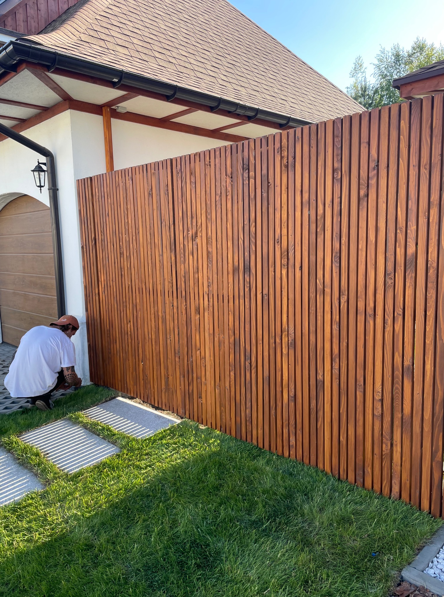 Montaż drewnianego ogrodzenia panelowego z pionowych desek. Widoczny fragment domu i trawnik z kamiennymi płytami. Fachowiec w trakcie pracy.