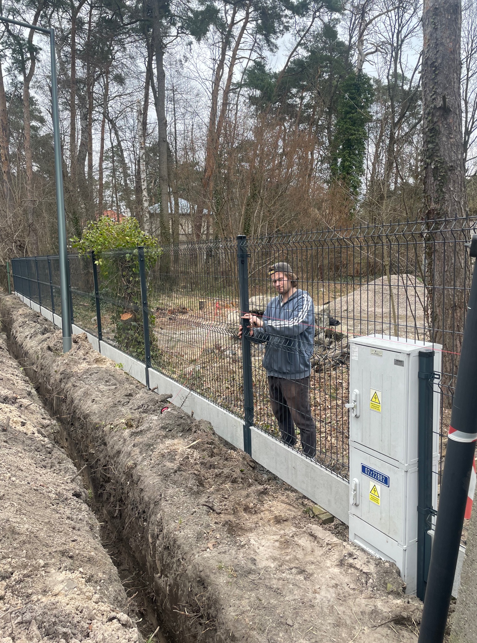 Montaż ogrodzenia panelowego z podmurówką betonową. Widoczny pracownik przy panelu, wykop pod fundament. Na słupie skrzynka elektryczna. Tło: drzewa.