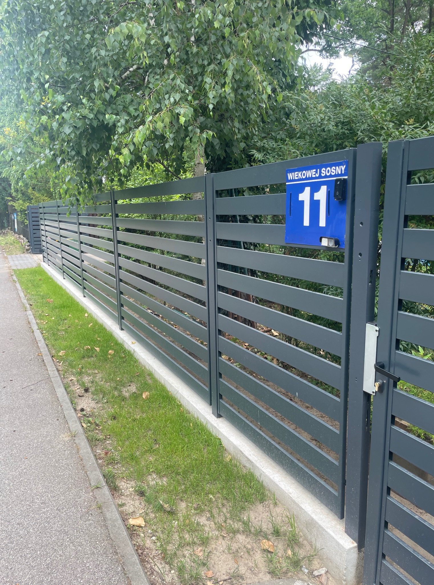 Nowoczesne, poziome ogrodzenie panelowe w kolorze grafitowym, z numerem posesji '11' na niebieskiej tabliczce. Widok z boku, z zielenią w tle.