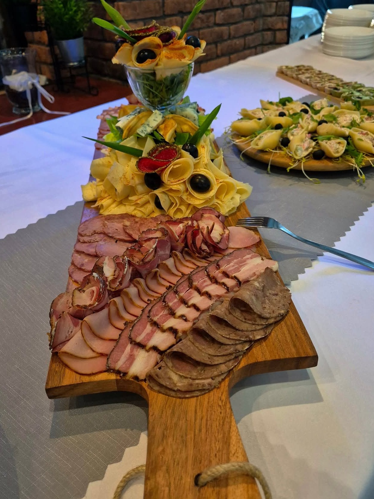 Dekoracyjna deska wędlin i serów z ozdobami z warzyw i owoców, część zastawy stołowej w tle. Elegancki catering na przyjęcie.
