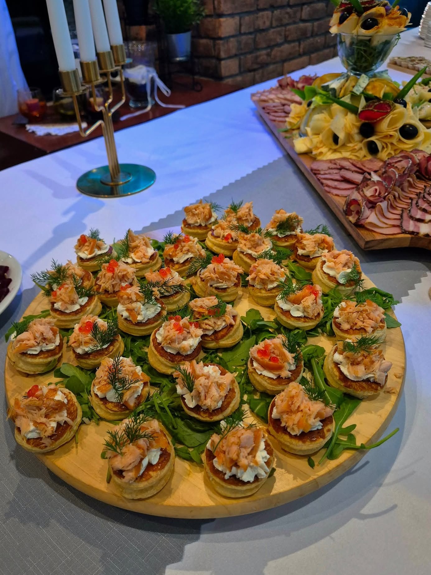 Elegancko podane mini tartaletki z łososiem, kremowym serem i koperkiem na drewnianej desce, część zastawionego stołu w tle. Dekoracja stołu na przyjęcie.