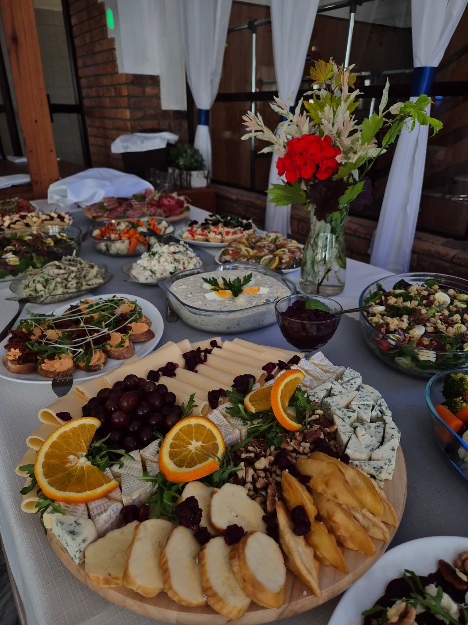 Bogato zastawiony stół cateringowy z różnorodnymi przekąskami: sery, sałatki, pieczywo, owoce i kwiaty w wazonie. Dekoracja z białych zasłon w tle.