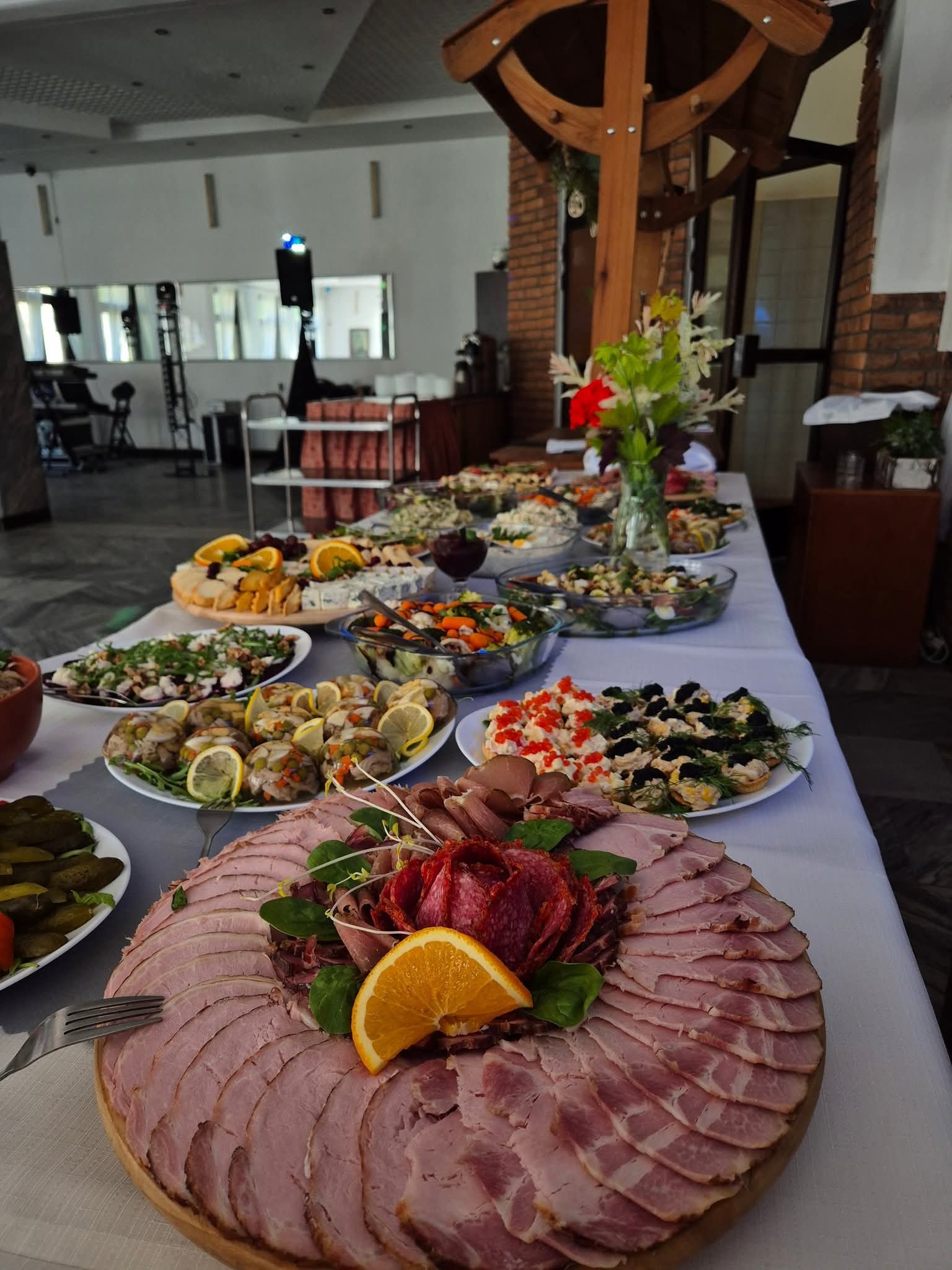 Stół bankietowy z różnorodnymi potrawami: wędliny, sałatki, sery i przekąski. Dekoracja z kwiatów i owoców. Widoczne elementy wyposażenia sali.