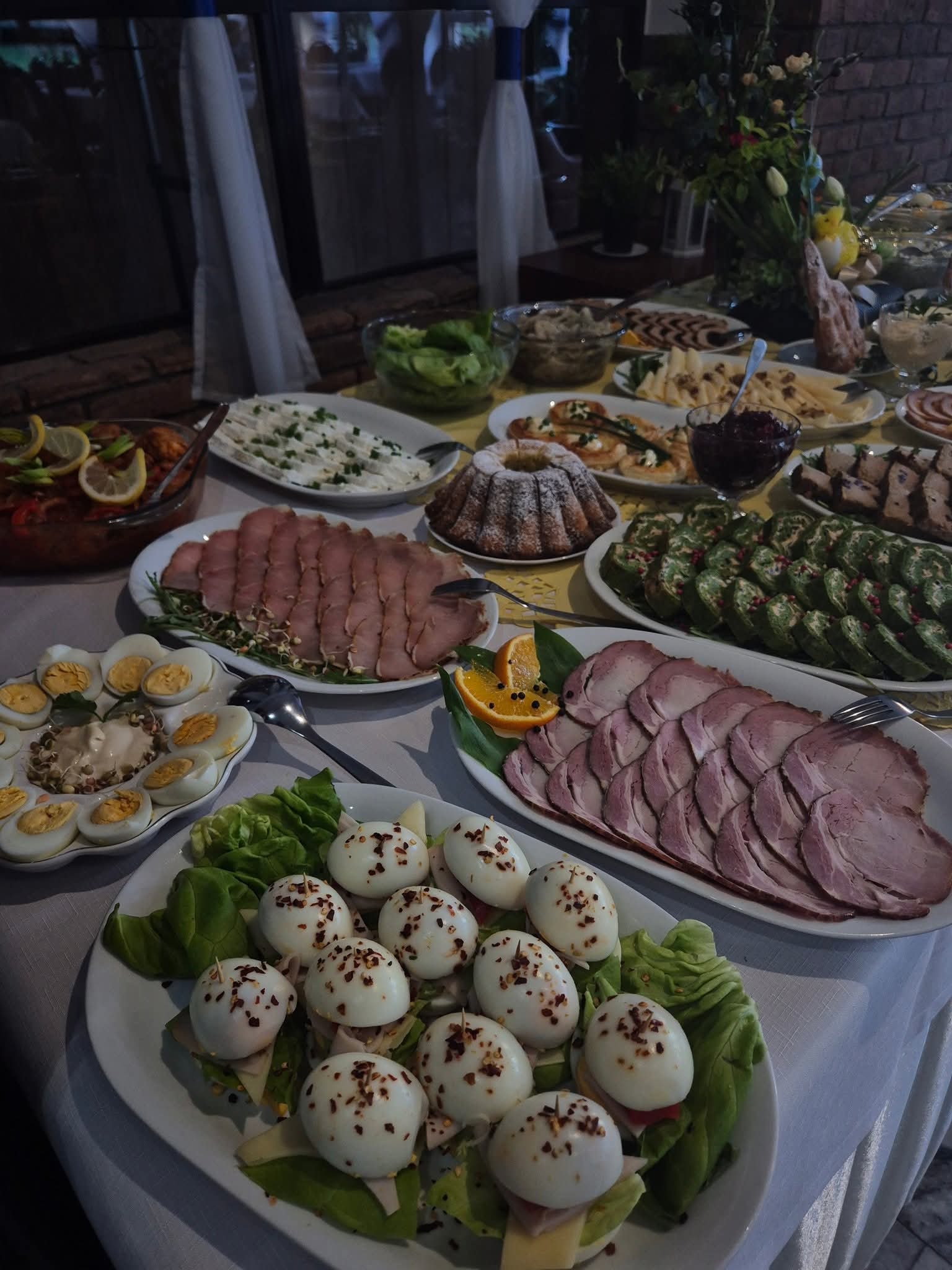 Bogato zastawiony stół z różnorodnymi daniami: wędliny, jajka, sałatki, ciasta. Dekoracja kwiatowa w tle. Idealne na przyjęcie okolicznościowe.