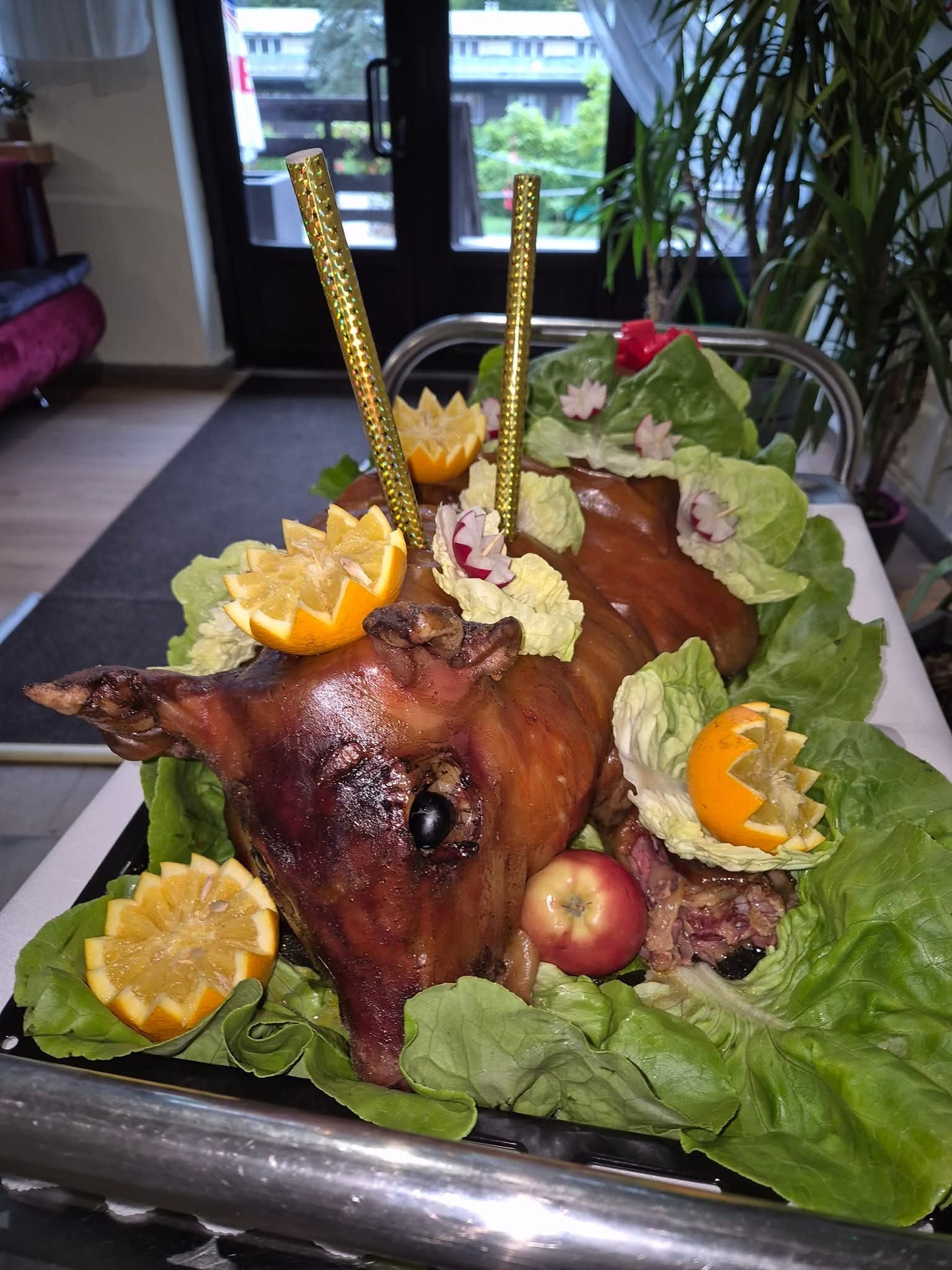 Prosię pieczone, dekorowane pomarańczami, jabłkiem i sałatą, umieszczone na metalowym wózku cateringowym z dwoma złotymi świeczkami.