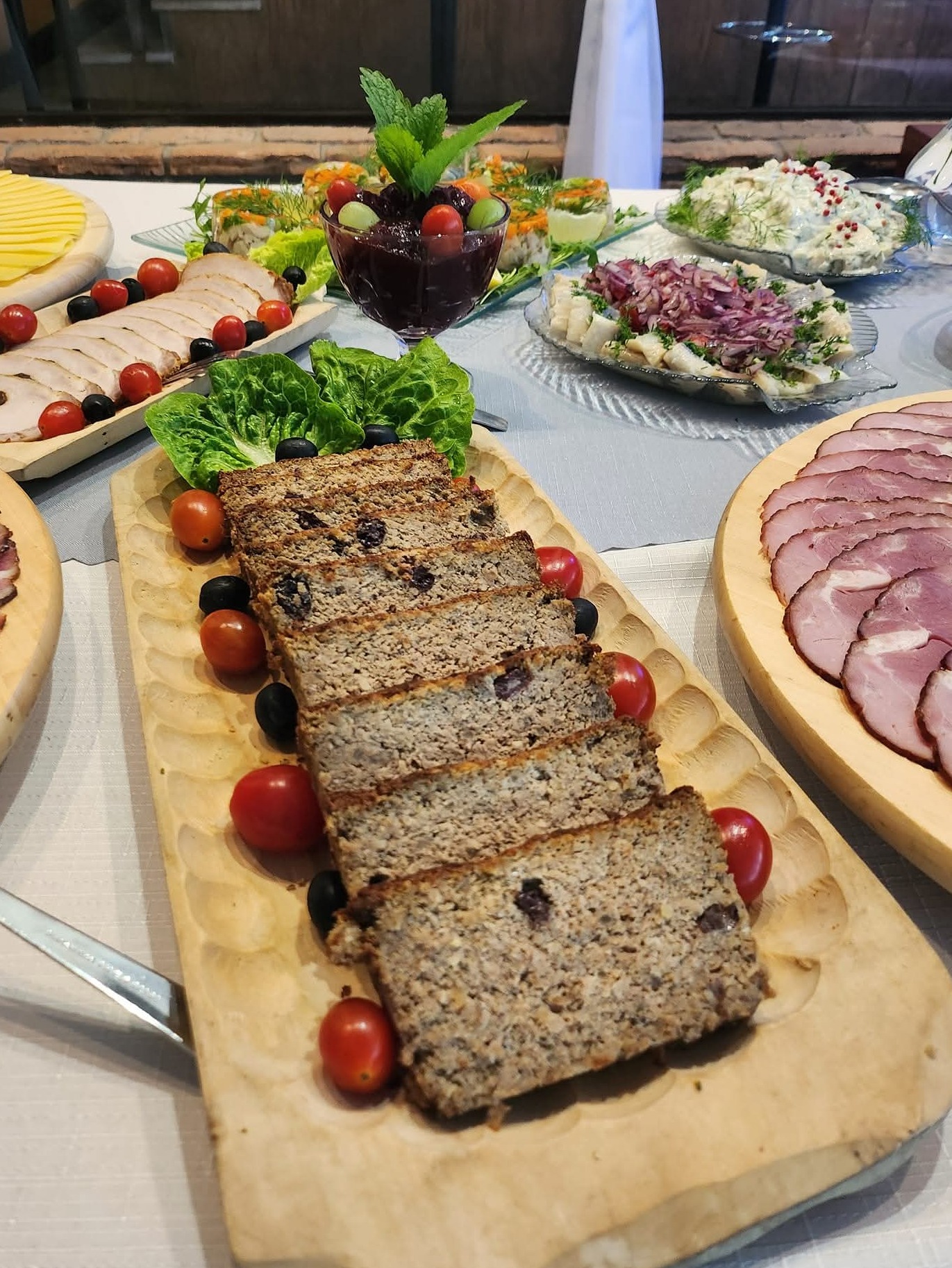 Stół z różnorodnymi potrawami cateringowymi: pasztet na drewnianej desce, wędliny, sałatki i dekoracje z warzyw. Ujęcie z bliska, ciepłe światło.