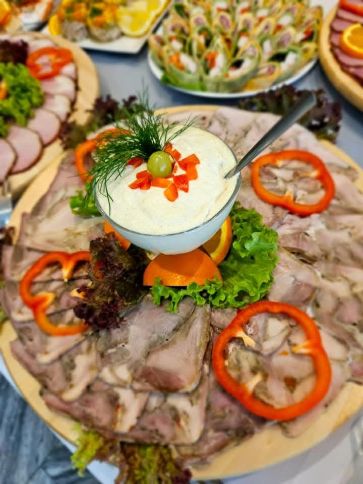 Dekoracyjna taca z wędlinami, papryką i sosem w miseczce, ozdobiona zieleniną. Widoczne inne tace z przekąskami w tle. Elegancki catering na przyjęcie.