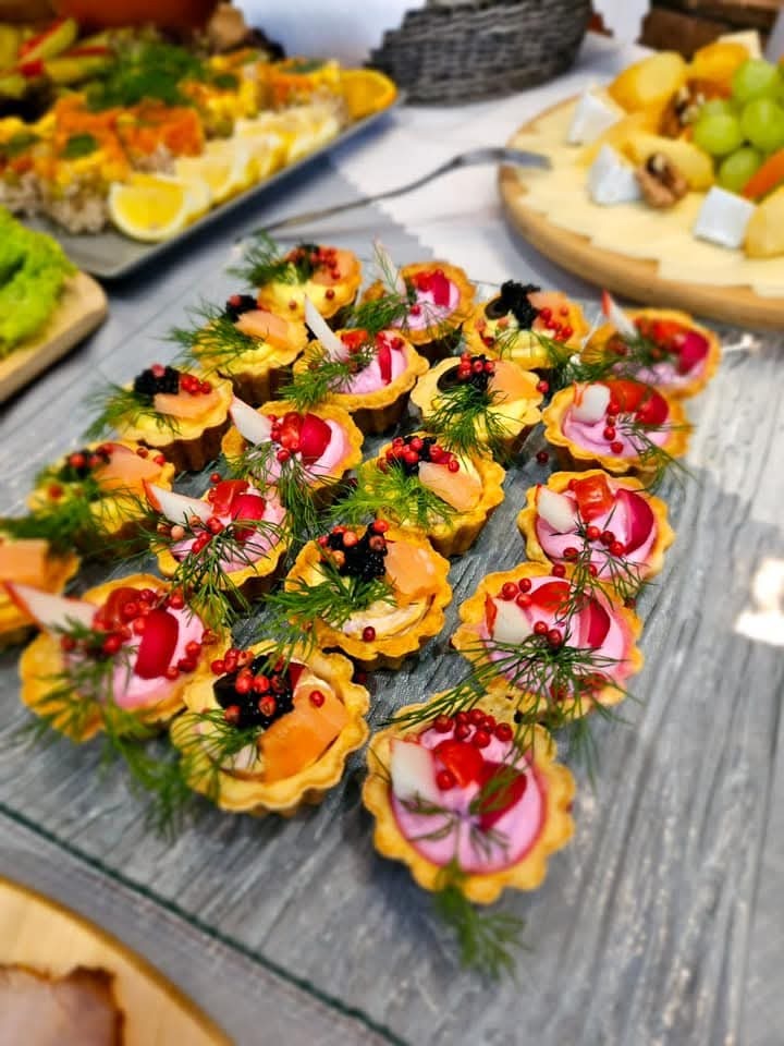 Eleganckie tartaletki z łososiem, kawiorem i kremowym nadzieniem, udekorowane koperkiem i czerwonym pieprzem, ułożone na szklanym talerzu. W tle inne przekąski.