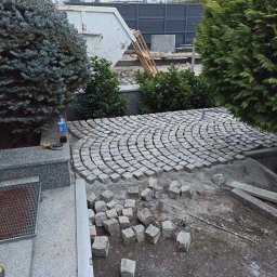 BVM Brykarstvo - Fragment układanej nawierzchni z kostki w łukowaty wzór, z widocznymi narzędziami i materiałami budowlanymi, otoczony zielenią i elementami infrastruktury.