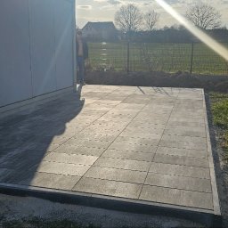 BVM Brykarstvo - Ułożona, szara nawierzchnia z obrzeżami przy budynku, z widocznym zagęszczarką w tle. Osoba częściowo widoczna przy ścianie budynku. Słoneczny dzień.