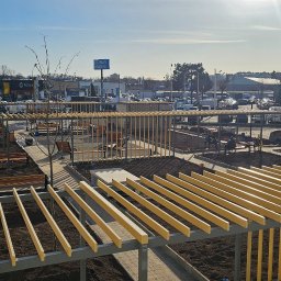 BVM Brykarstvo - Widok z góry na plac z alejkami z kostki, drewnianymi pergolami i ławkami. W tle budynki i parking. Prace ziemne w toku, widoczny sprzęt budowlany.