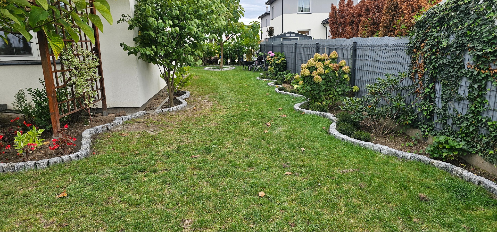 Ogród z trawnikiem i rabatami kwiatowymi, obrzeża z szarej kostki, falista linia. W tle dom i ogrodzenie z roślinami. Widok z perspektywy trawnika.