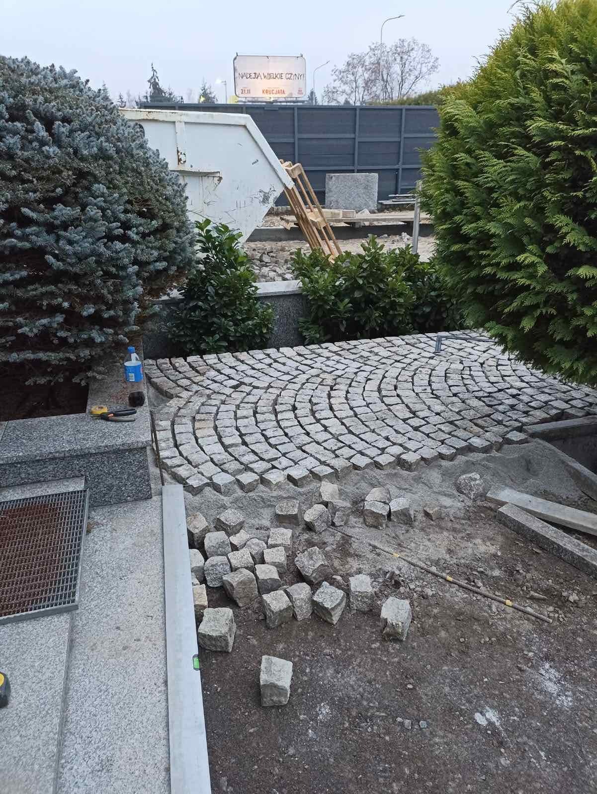 Fragment układanej nawierzchni z kostki w łukowaty wzór, z widocznymi narzędziami i materiałami budowlanymi, otoczony zielenią i elementami infrastruktury.