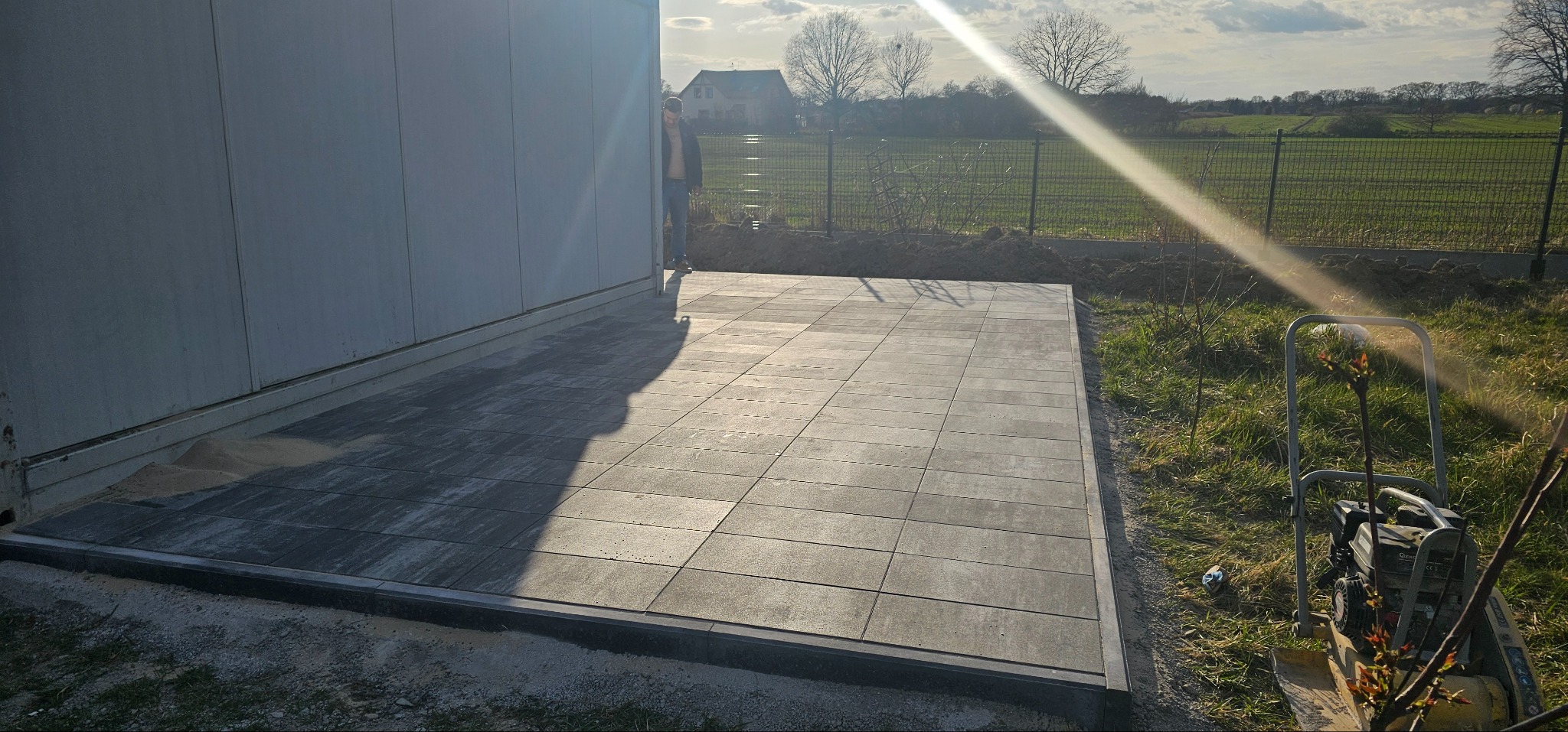 Ułożona, szara nawierzchnia z obrzeżami przy budynku, z widocznym zagęszczarką w tle. Osoba częściowo widoczna przy ścianie budynku. Słoneczny dzień.