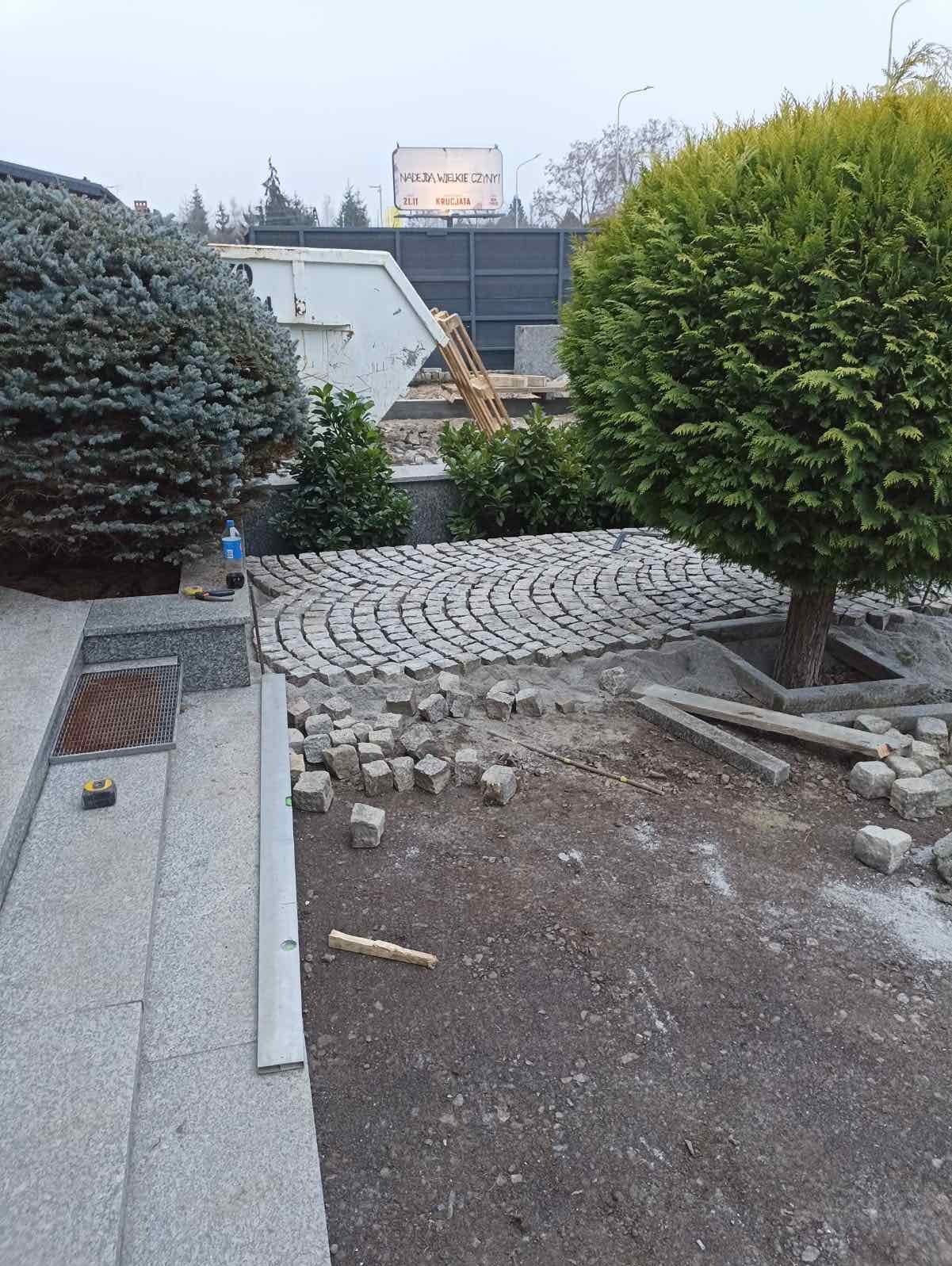 Fragment ogrodu w trakcie prac: ułożona część okrągłej ścieżki z kostki granitowej, obok leżą nieułożone kostki, widoczne narzędzia i roślinność. Prace brukarskie w toku.