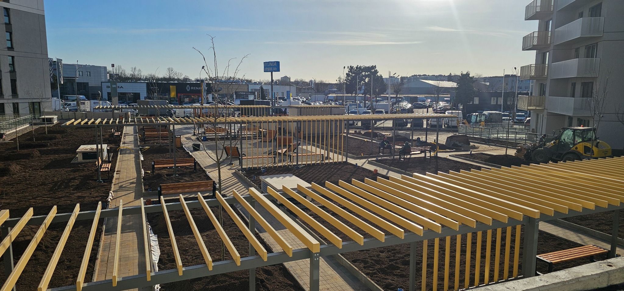 Widok z góry na plac z alejkami z kostki, drewnianymi pergolami i ławkami. W tle budynki i parking. Prace ziemne w toku, widoczny sprzęt budowlany.