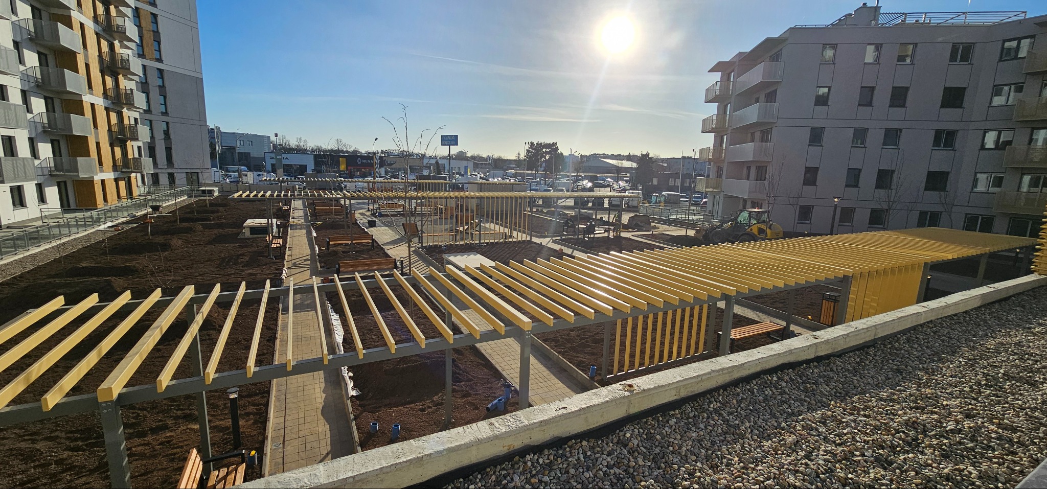 Nowoczesny park osiedlowy z pergolami i ścieżkami z kostki. W tle budynki mieszkalne i plac budowy z koparką. Słoneczny dzień, widok z góry.