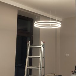 ROBO-TECHNICA SPÓŁKA Z OGRANICZONĄ ODPOWIEDZIALNOŚCIĄ - Nowoczesna, okrągła lampa LED zamontowana na suficie. Pod nią rozstawiona drabina aluminiowa, a obok widoczne narzędzia i stanowisko pracy. Świeże, jasne wnętrze.