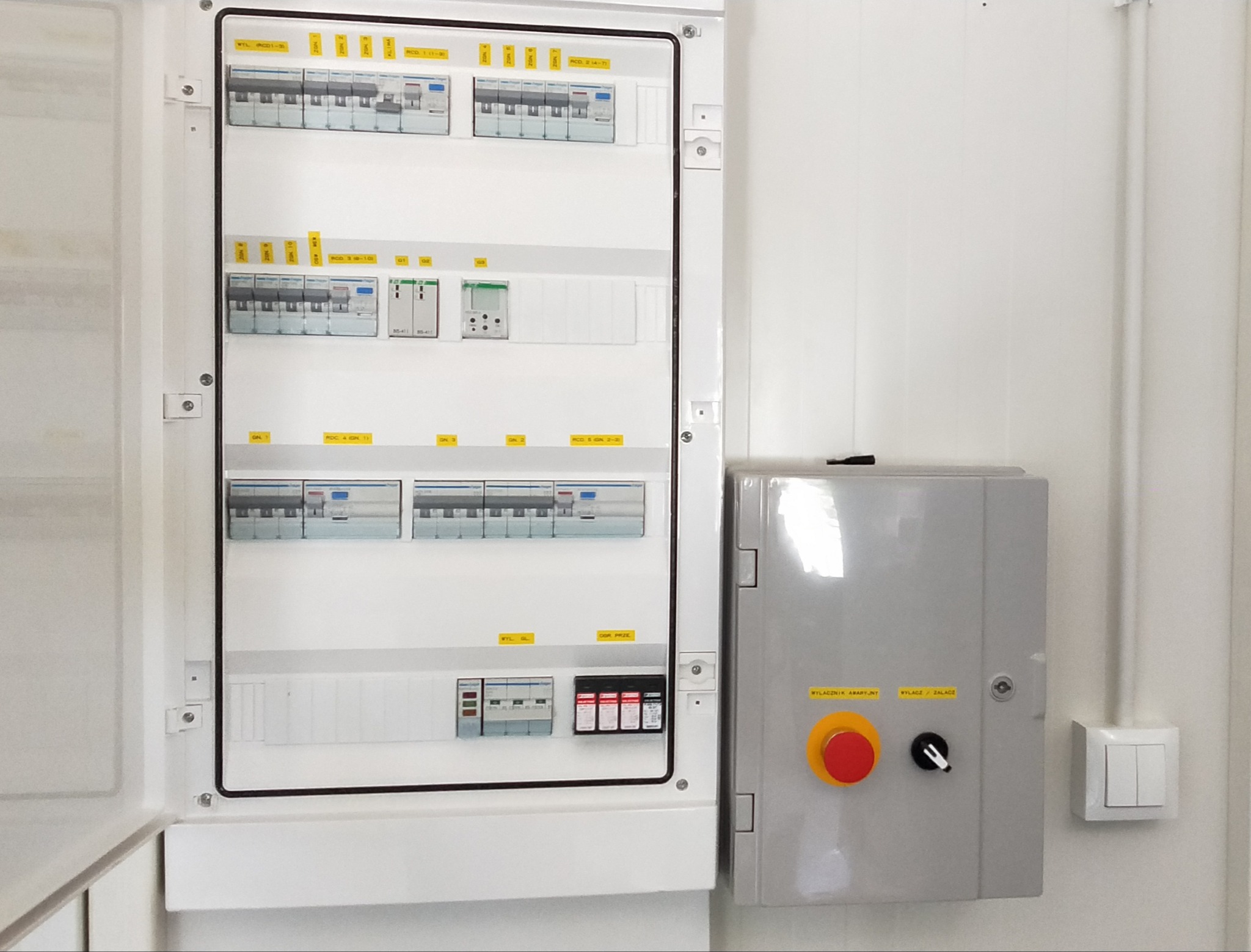 Otwarta, biała skrzynka rozdzielcza z bezpiecznikami i oznaczeniami na ścianie. Obok szara skrzynka z przyciskiem awaryjnym i wyłącznikiem. Widoczny fragment instalacji elektrycznej.