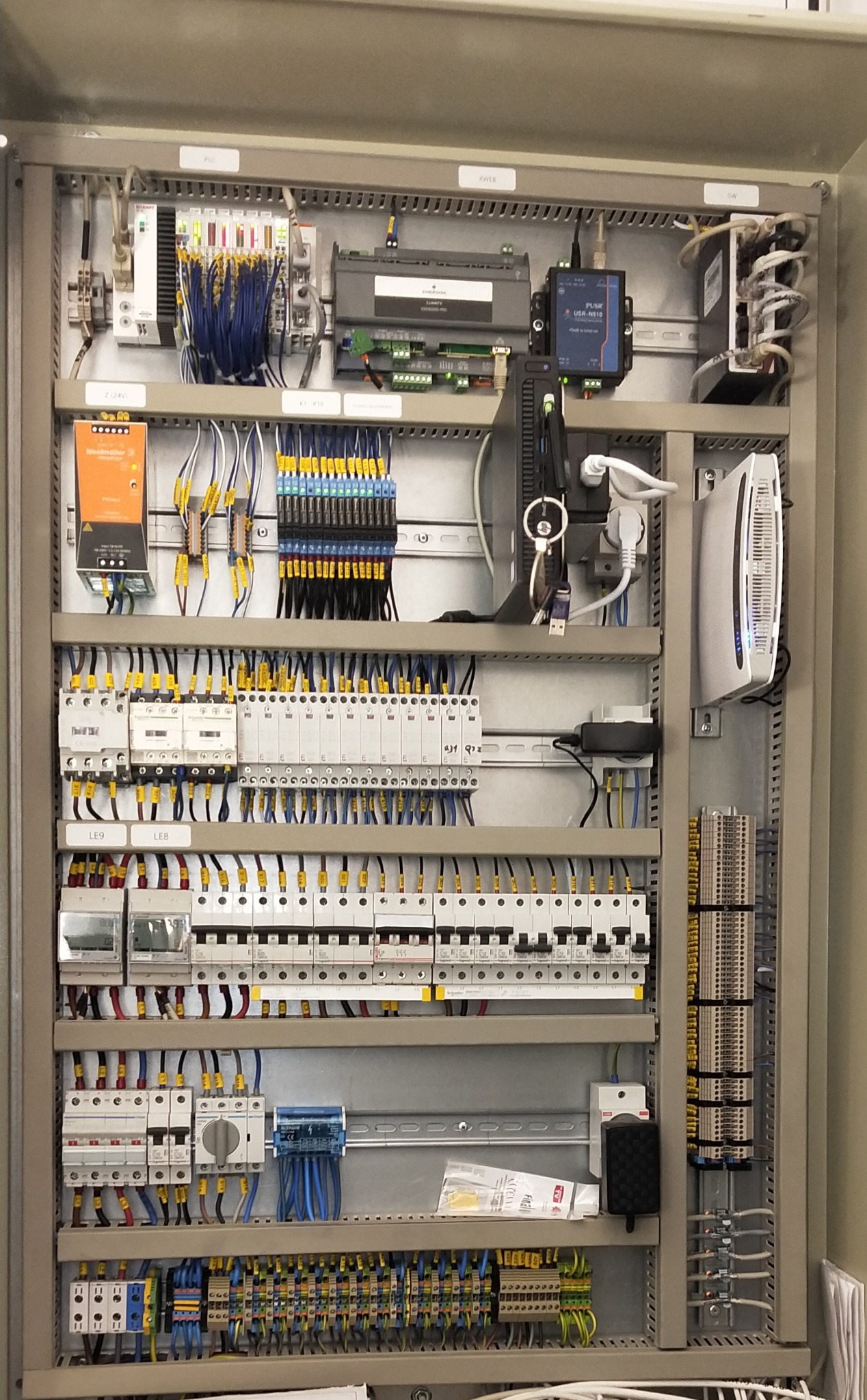 Rozdzielnica elektryczna z licznymi przewodami, bezpiecznikami i urządzeniami sterującymi. Widoczne kable w różnych kolorach, moduły PLC i elementy automatyki przemysłowej.