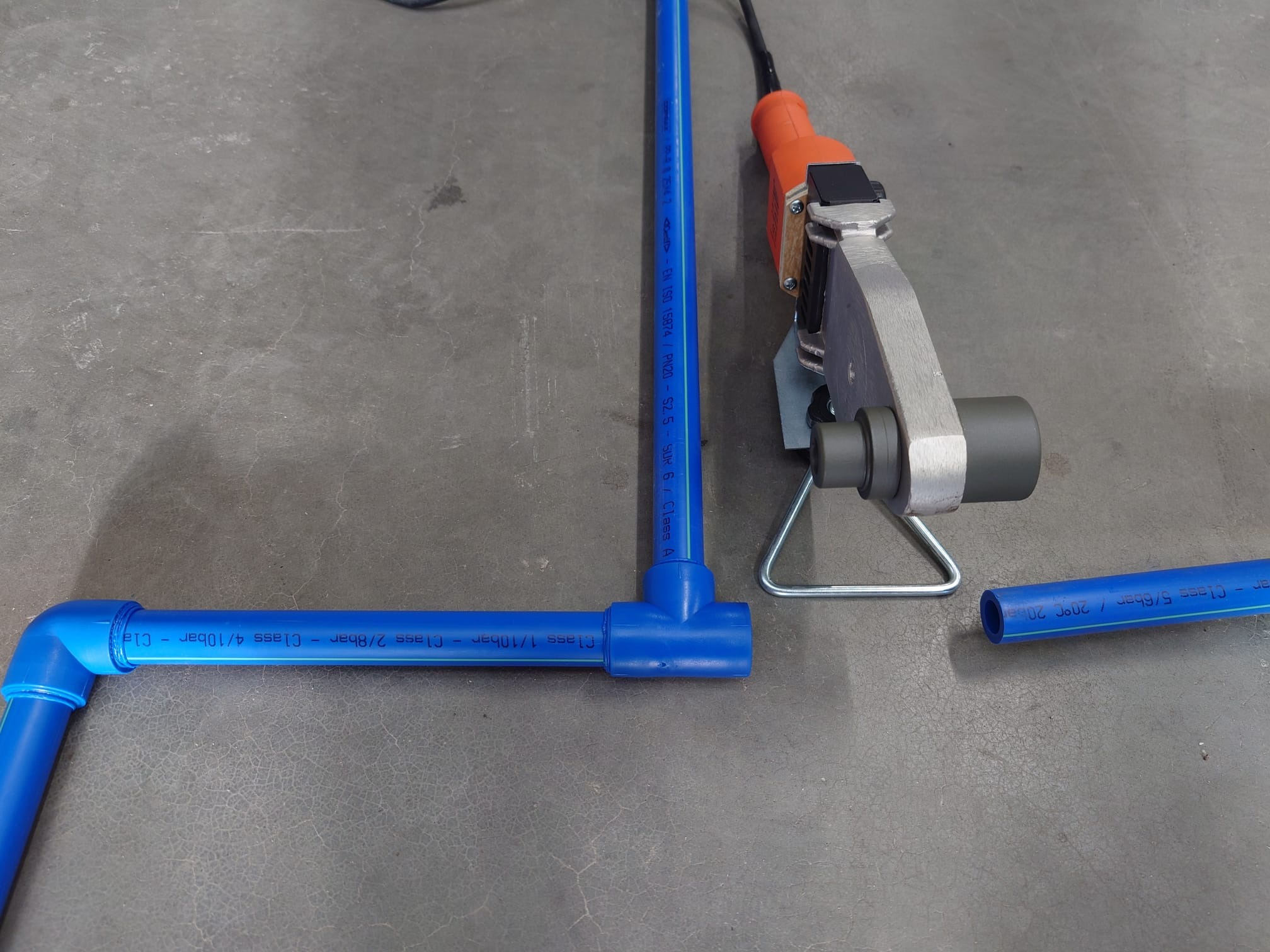 Niebieskie rury PP-R z kształtkami i zgrzewarką na szarym tle. Elementy instalacji wodnej przed montażem, widoczne oznaczenia ciśnienia i temperatury na rurach.