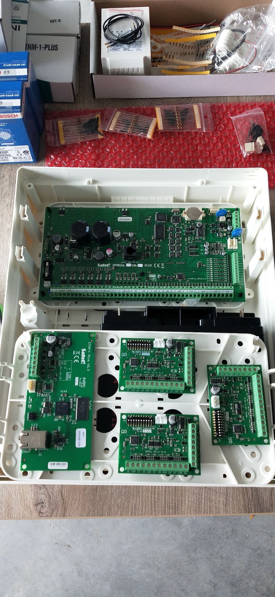 Otwarta obudowa centrali alarmowej Satel Integra 128 Plus z modułami rozszerzeń ETHM-1 Plus i INT-E, widoczne płytki drukowane i elementy elektroniczne. Akcesoria w tle.