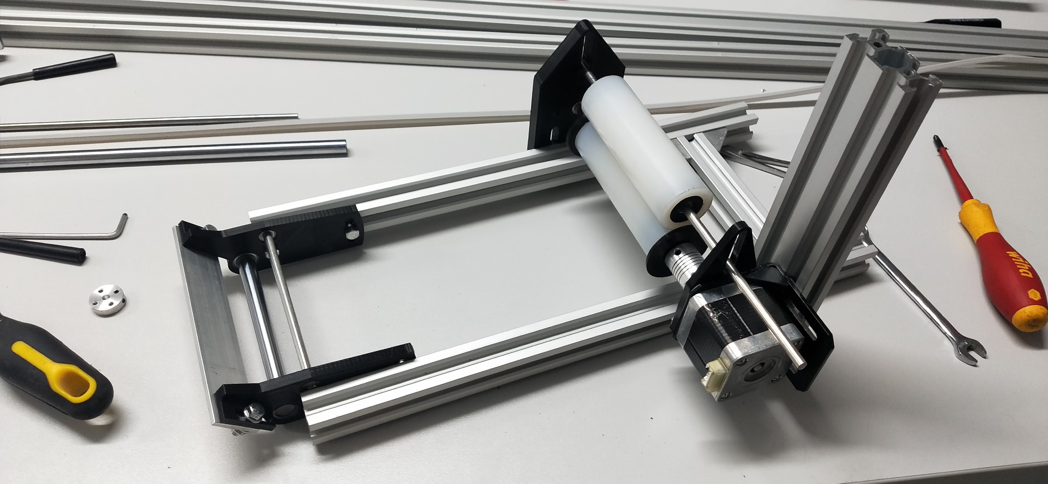 Konstrukcja z profili aluminiowych, silnika krokowego i rolek, w trakcie montażu. Widoczne narzędzia: śrubokręt Wiha i klucz płaski. Elementy na białym stole.