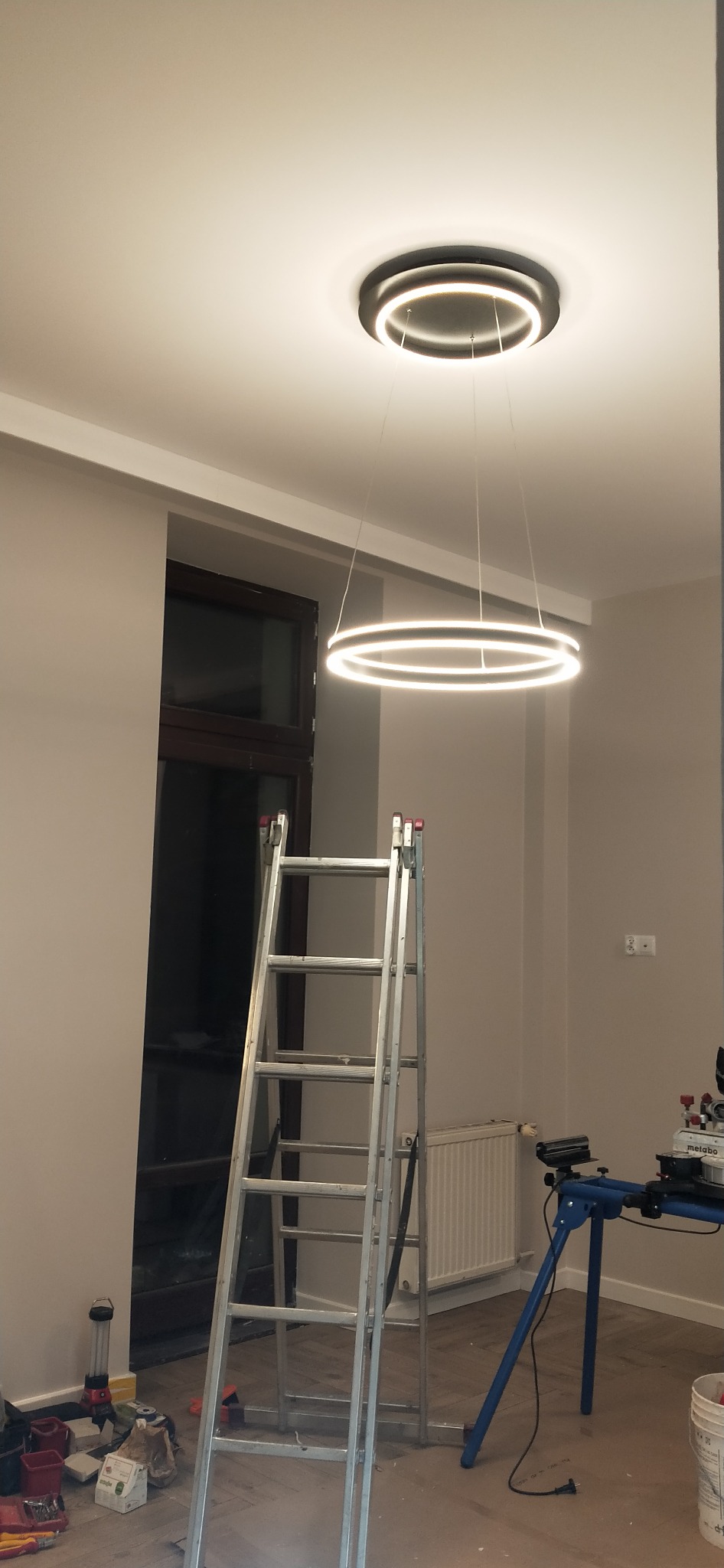 Nowoczesna, okrągła lampa LED zamontowana na suficie. Pod nią rozstawiona drabina aluminiowa, a obok widoczne narzędzia i stanowisko pracy. Świeże, jasne wnętrze.