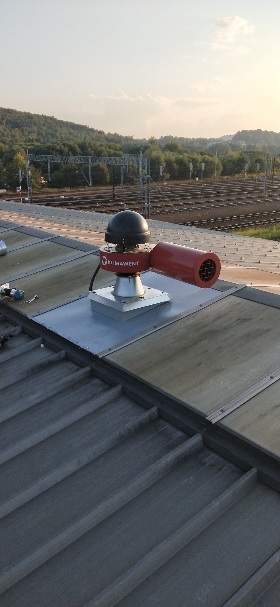 Zamontowany na dachu wentylator przemysłowy marki Klimawent, z widocznymi elementami mocującymi i narzędziami. W tle zieleń i tory kolejowe.