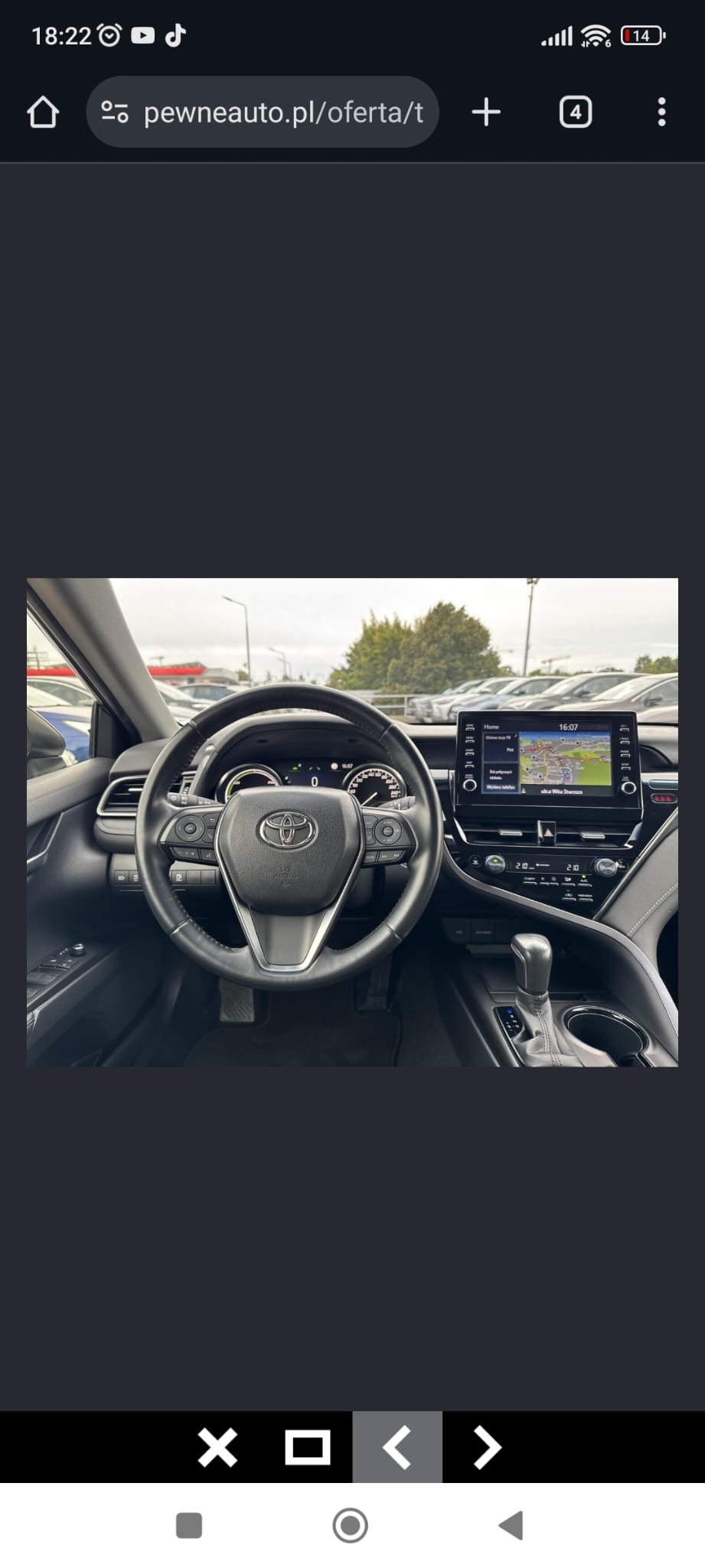 Wnętrze samochodu Toyota Camry z widocznym kokpitem, kierownicą z logo, nawigacją GPS i automatyczną skrzynią biegów. Na zewnątrz parking z innymi autami.