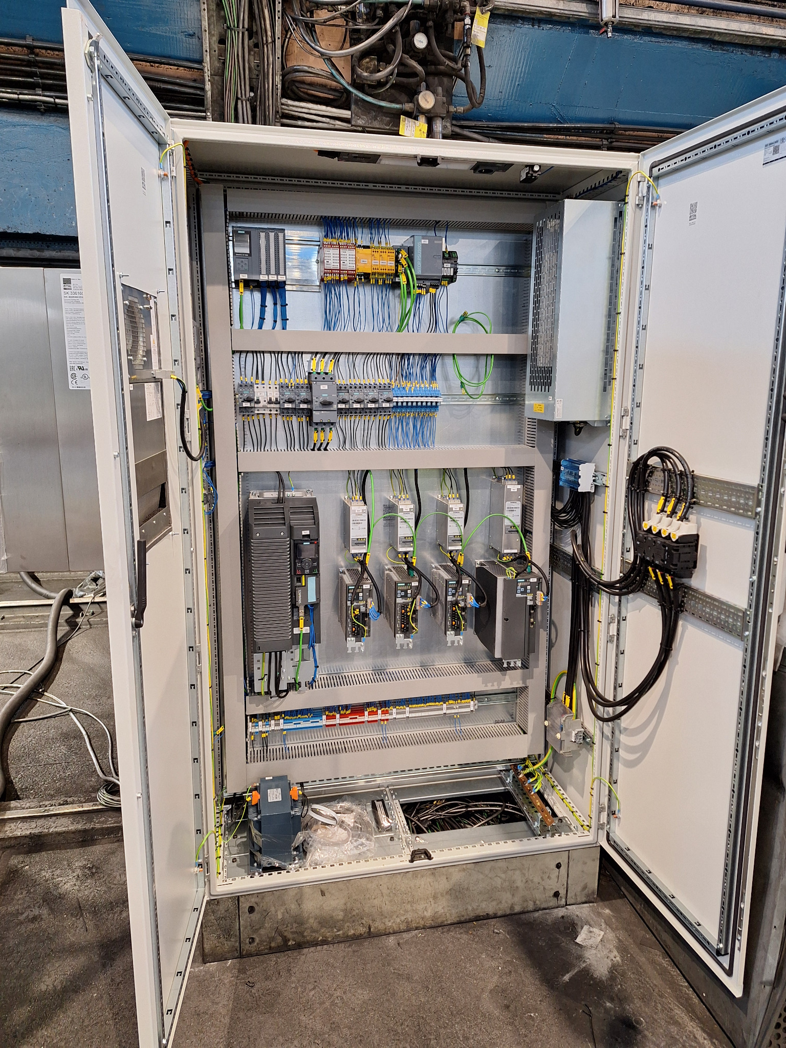 Otwarta szafa sterownicza z widocznymi elementami elektrycznymi: sterowniki, kable, złączki. Precyzyjny montaż w szarej obudowie. Widoczne przewody w różnych kolorach.