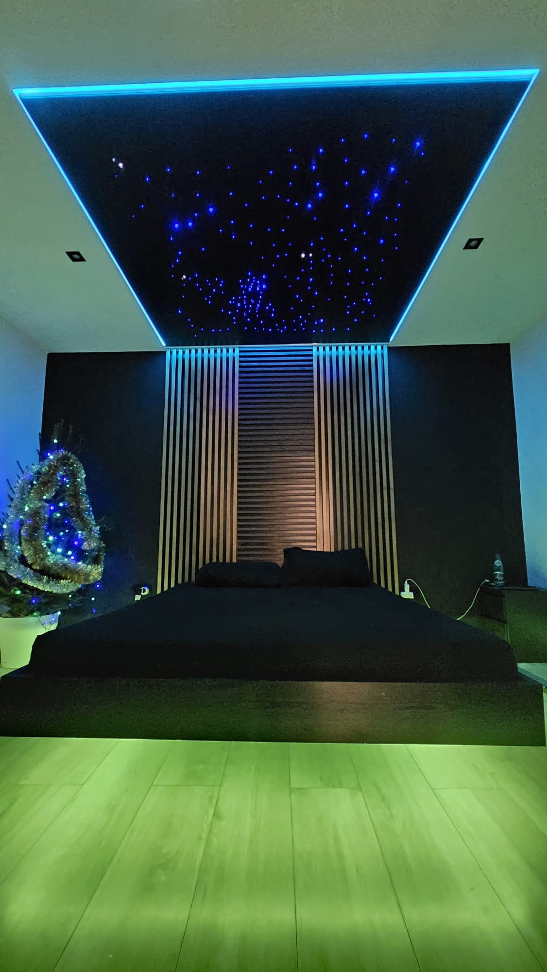Sypialnia z czarnym łóżkiem, podświetlaną podłogą i sufitem napinanym imitującym nocne niebo z gwiazdami, obok choinka. Nowoczesny design.