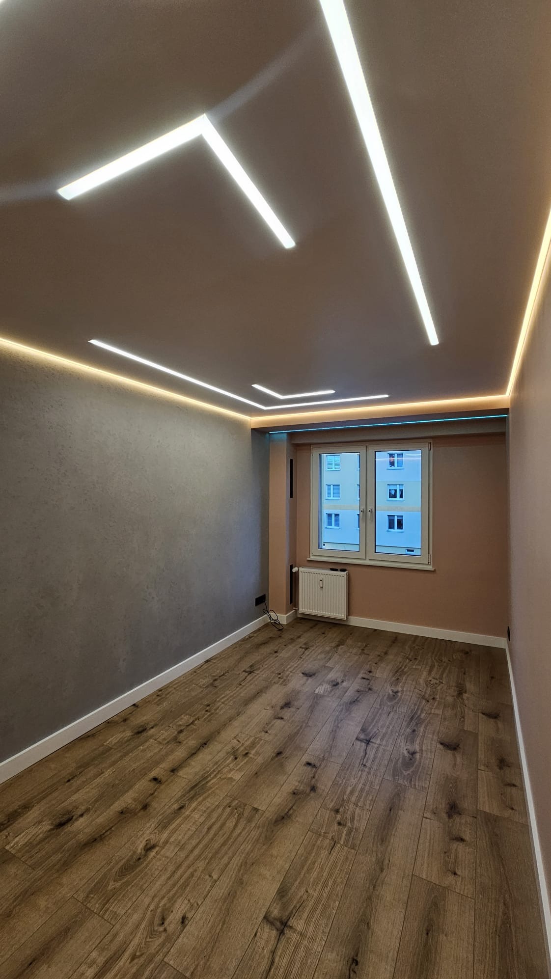 Pokój po remoncie z drewnianą podłogą, szarą ścianą i oświetleniem LED na suficie. Widok na okno z grzejnikiem. Nowoczesny design wnętrza.