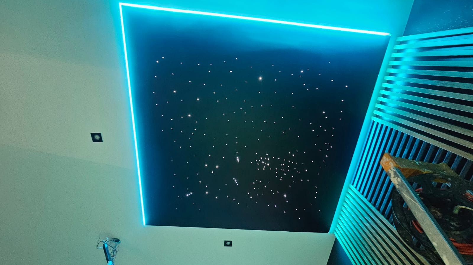 Nastrojowy sufit napinany z efektem gwiazd, podświetlony neonowym obramowaniem. Fragment dekoracyjnej ściany z listew i drabina malarska w prawym dolnym rogu.