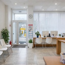 EKSPERT MAŁGORZATA ZAJKOWSKA - Jasne wnętrze biura z recepcją, białymi krzesłami i roślinami doniczkowymi. Widoczne monitory, rolety pionowe i przeszklone drzwi wejściowe.